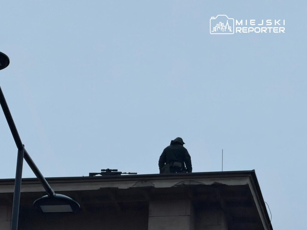 Funkcjonariusz w czarnym ubraniu stoi na dachu budynku, z bronią snajperską obok.