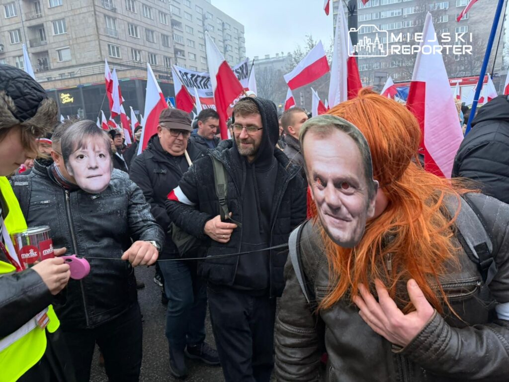 Grupa osób w maskach z wizerunkami polityków uczestniczy w demonstracji, trzymając flagi narodowe.