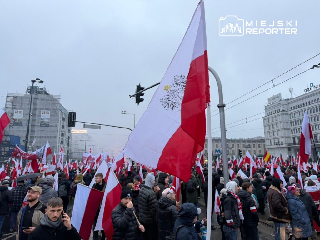 Tłum ludzi z flagami Polski na ulicy w szarym, pochmurnym dniu, otoczeni nowoczesnymi budynkami.