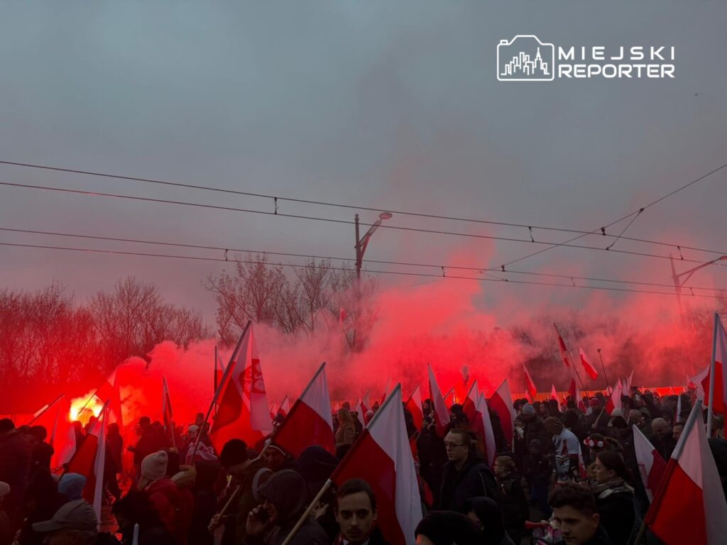 Tłum ludzi z flagami narodowymi i czerwonymi racami na ulicy, w tle dym i szare niebo.