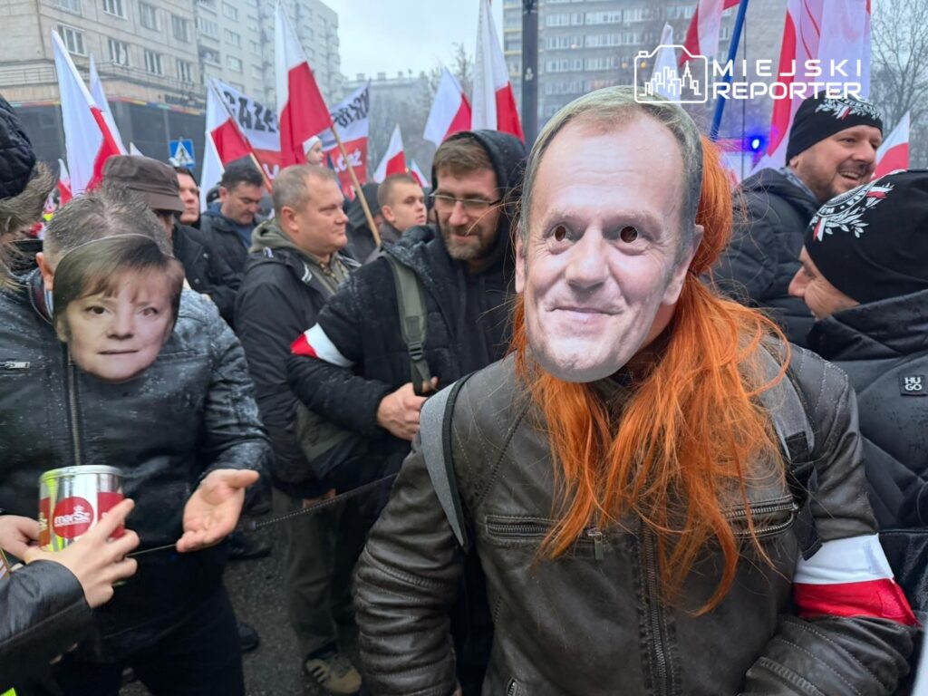 Grupa ludzi na demonstracji nosi maski z wizerunkami znanych postaci, trzymając flagi.