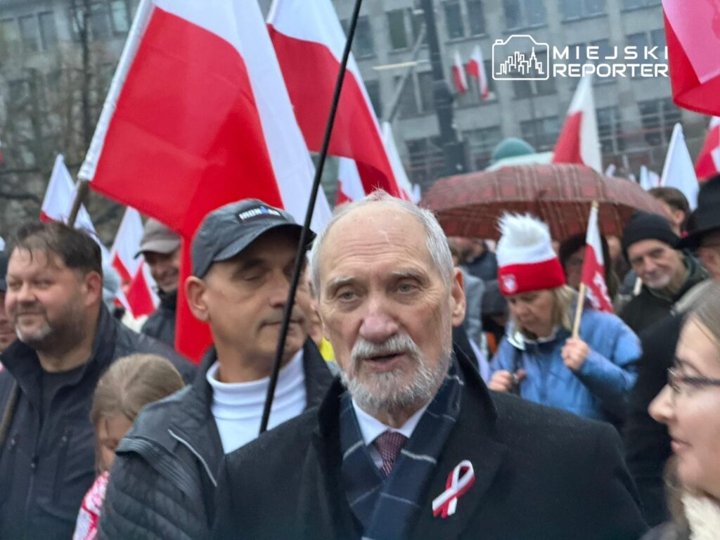 Mężczyzna z siwą brodą i w ciemnym płaszczu rozmawia z uczestnikami marszu, w tle powiewają polskie flagi.