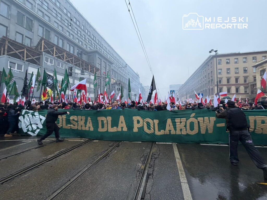 Tłum ludzi z flagami narodowymi i transparentem "POLSKA DLA POLAKÓW" przechodzi przez ulicę w miejskim otoczeniu.