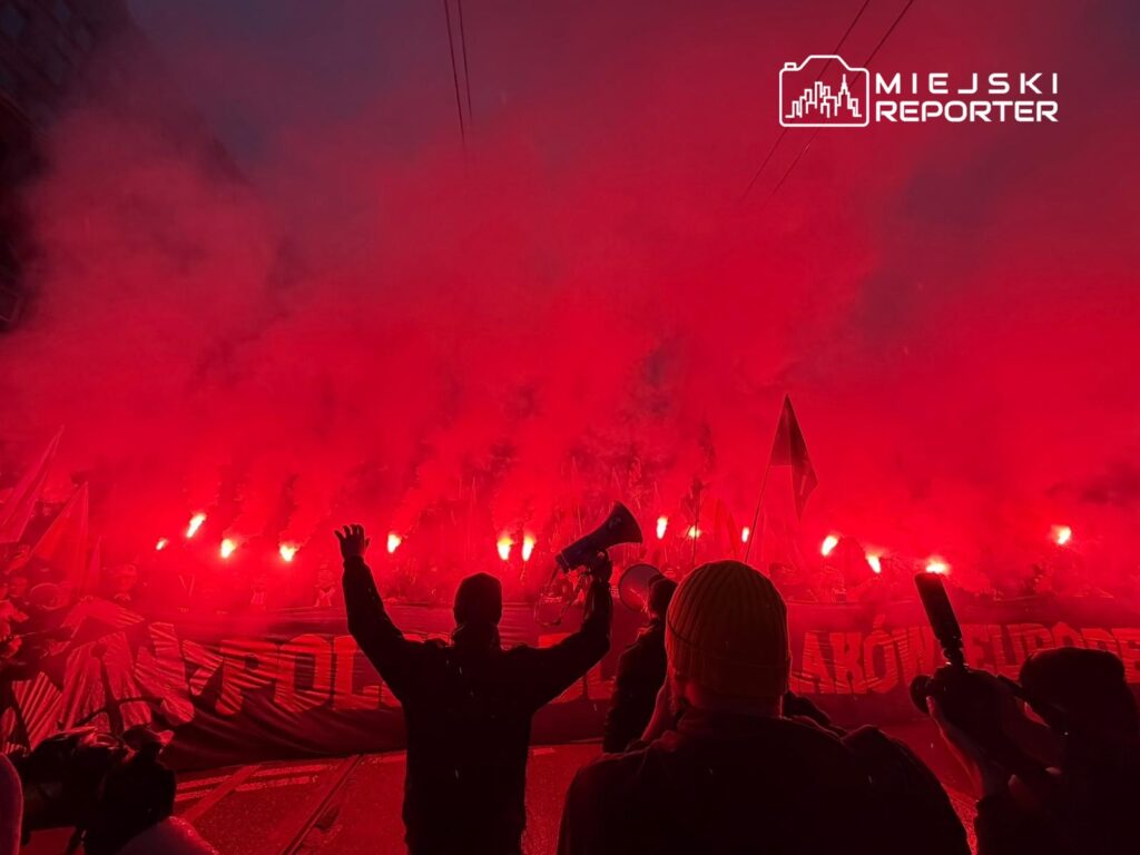 Tłum ludzi z flagami i transparentami, otoczony czerwonym dymem z rac, podczas manifestacji.
