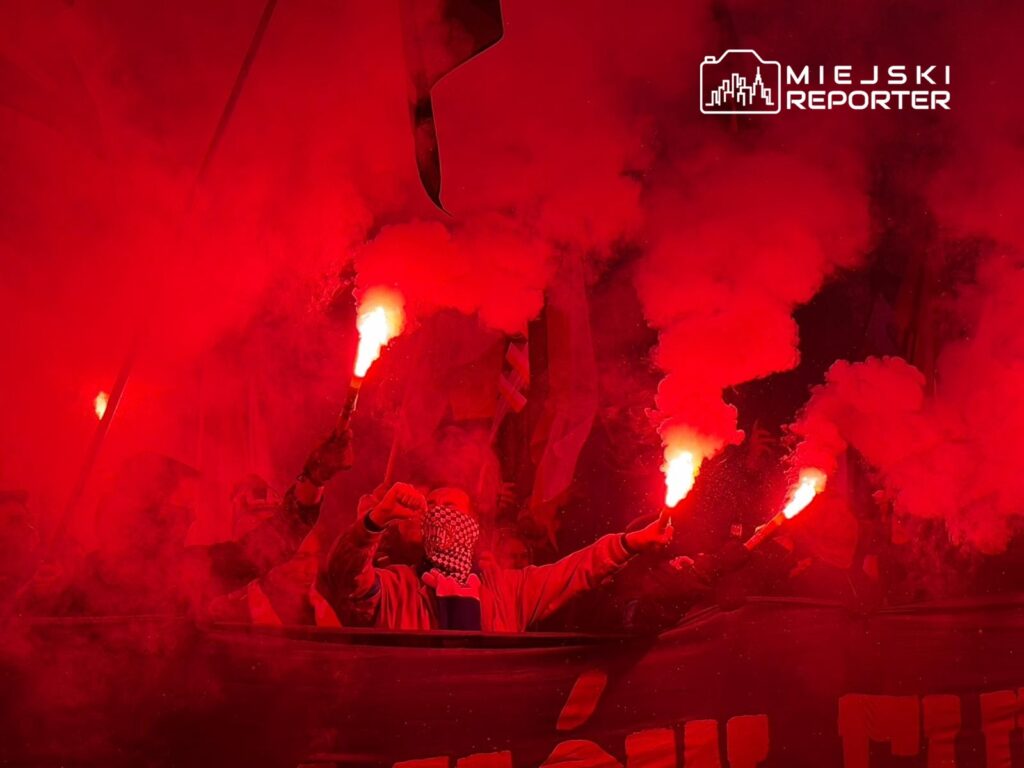 Grupa ludzi z flagami i racami świetlnymi w czerwonym dymie, manifestująca w nocy.