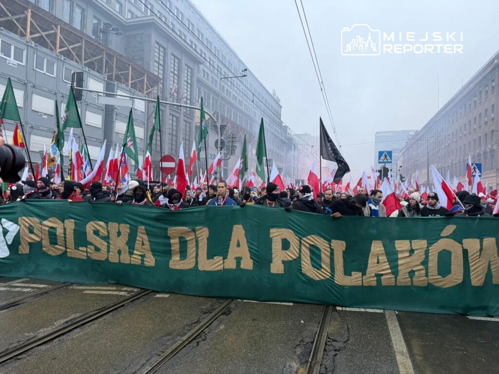 Tłum ludzi z flagami wznoszą transparent "POLSKA DLA POLAKÓW" na ulicy w miejskim otoczeniu.