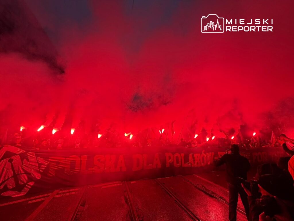 Grupa ludzi z flagami i transparentem "POLSKA DLA POLAKÓW" w tłumie, otoczona czerwoną mgłą dymu.