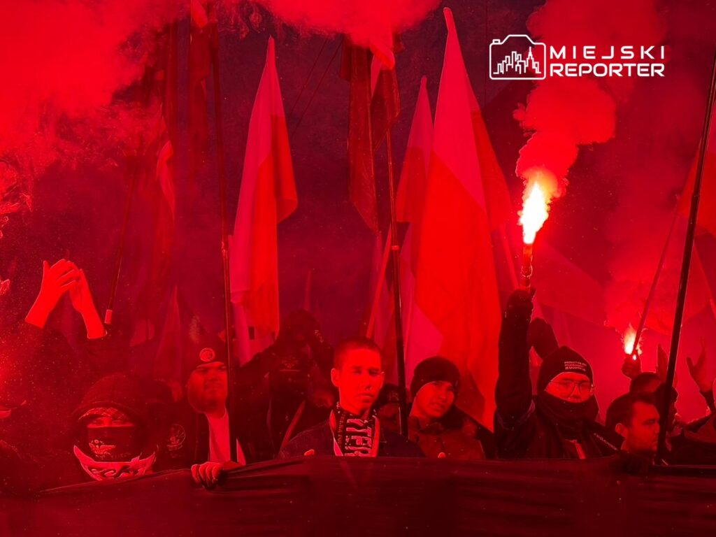 Grupa ludzi z flagami w tle, trzymająca race, tworzy intensywną atmosferę w czerwonym świetle.