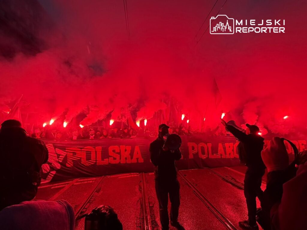 Tłum manifestujących z flagami i racami, wypełniających ulicę czerwoną mgłą dymu, z transparentem "POLSKA".
