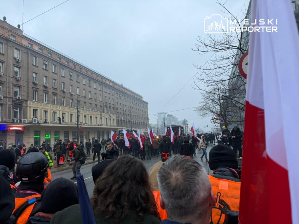 Grupa ludzi z flagami narodowymi maszeruje ulicą w mieście, w tle widoczny szary budynek.