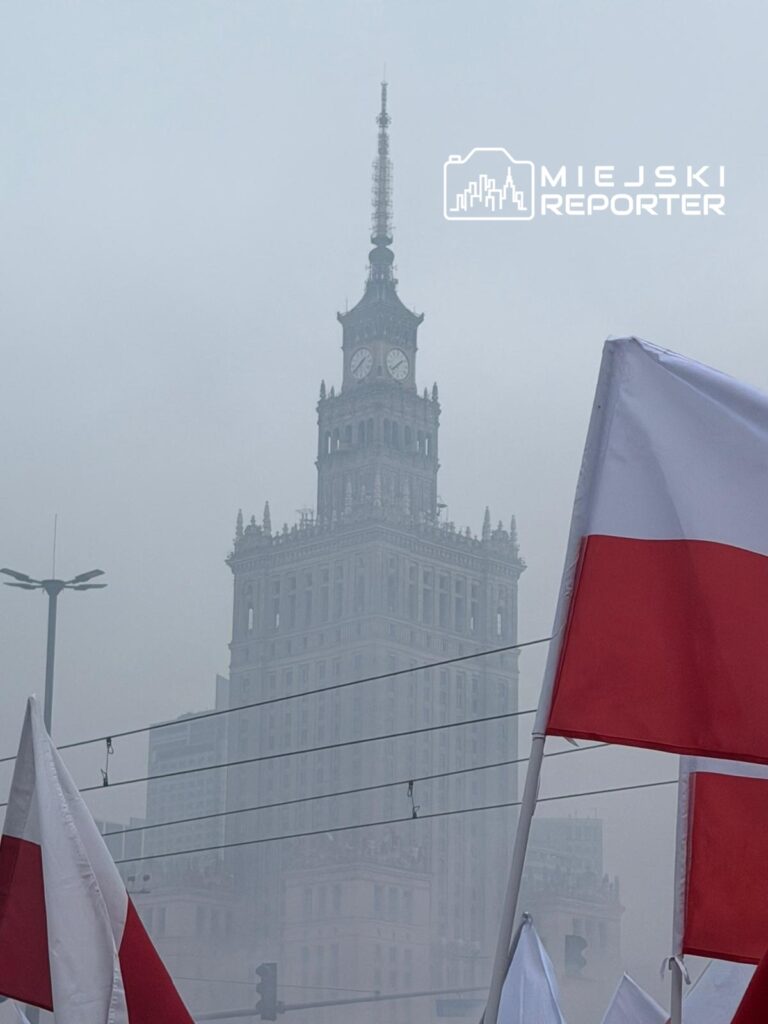 W tle widoczny zarys wieżowca z zegarem, na pierwszym planie powiewające flagi w kolorach białym i czerwonym.