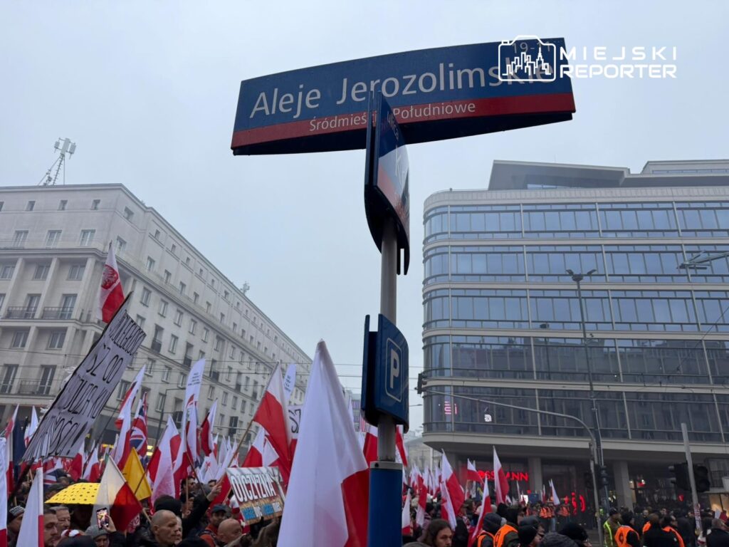 Tłum ludzi z flagami narodowymi i transparentami protestuje na ulicy, obok nowoczesnego budynku.