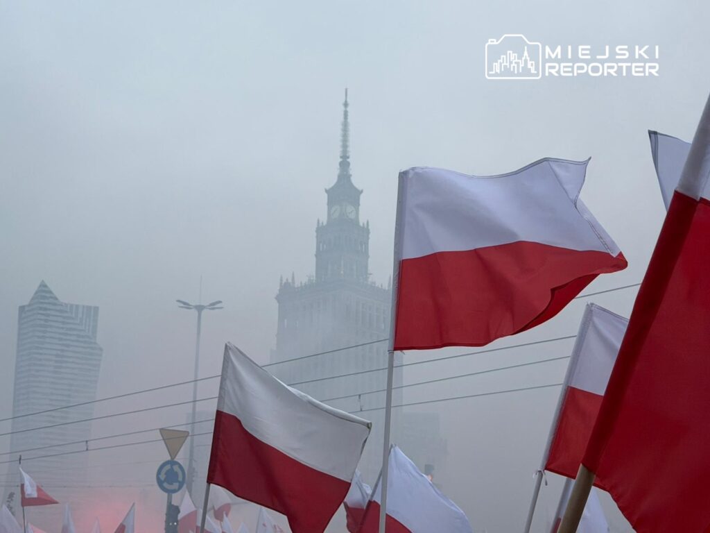 Grupa ludzi trzymająca flagi Polski, z widocznym w tle wieżowcem w mglistym otoczeniu.
