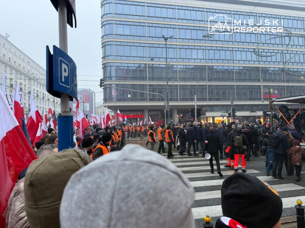 Tłum ludzi z flagami Polski gromadzi się na ulicy, a wśród nich widać osoby w pomarańczowych kamizelkach.