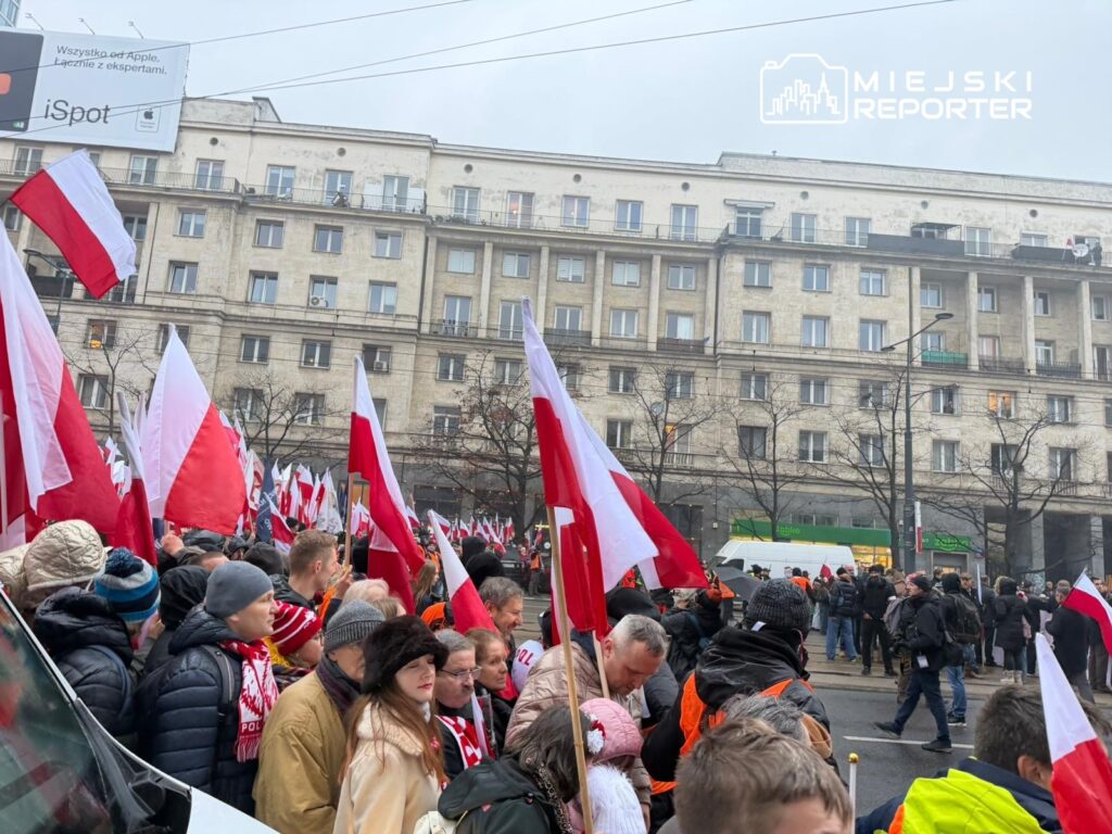 Tłum ludzi z flagami w barwach narodowych zgromadzony na ulicy w miejskim otoczeniu.