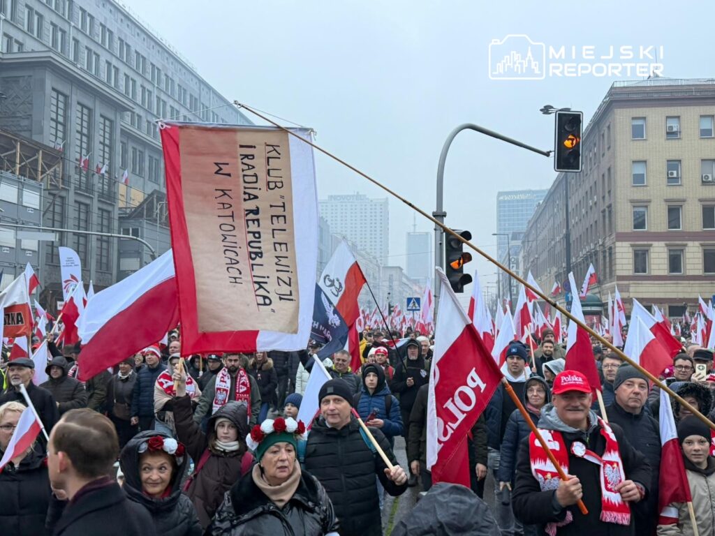 Tłum ludzi z flagami Polski uczestniczy w marszu, trzymając transparent z napisem "KLUB 'TELEWIZJI' IRADIA REPUBLIKA W KATOWICACH".