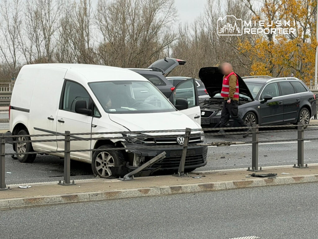 Biały samochód dostawczy z uszkodzoną przednią częścią stoi na jezdni, obok mężczyzna w odblaskowej kamizelce sprawdza uszkodzenia innego pojazdu.