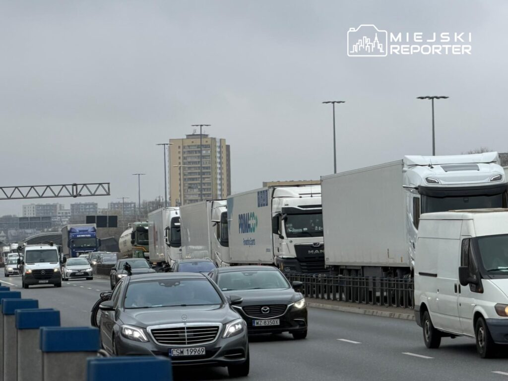 Ruch samochodowy na drodze z dużą liczbą ciężarówek i osobówek w miejskim otoczeniu. W tle widać wysokie budynki.