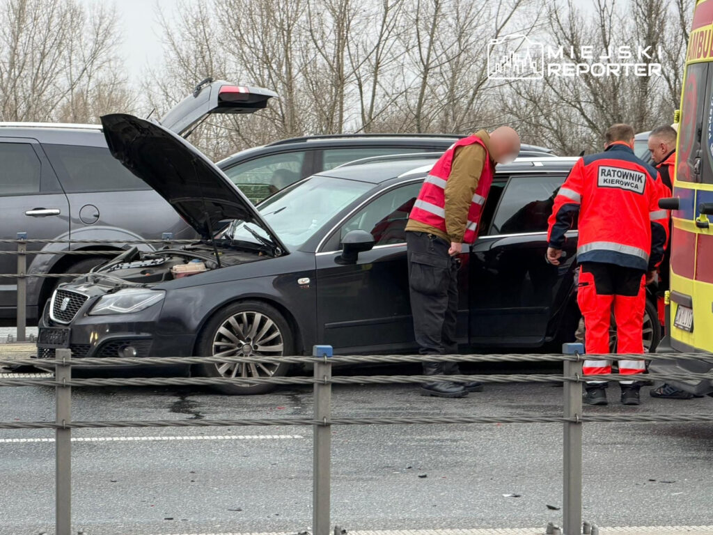 Mężczyzna w czerwonym stroju ratowniczym sprawdza silnik samochodu z otwartą maską na drodze.