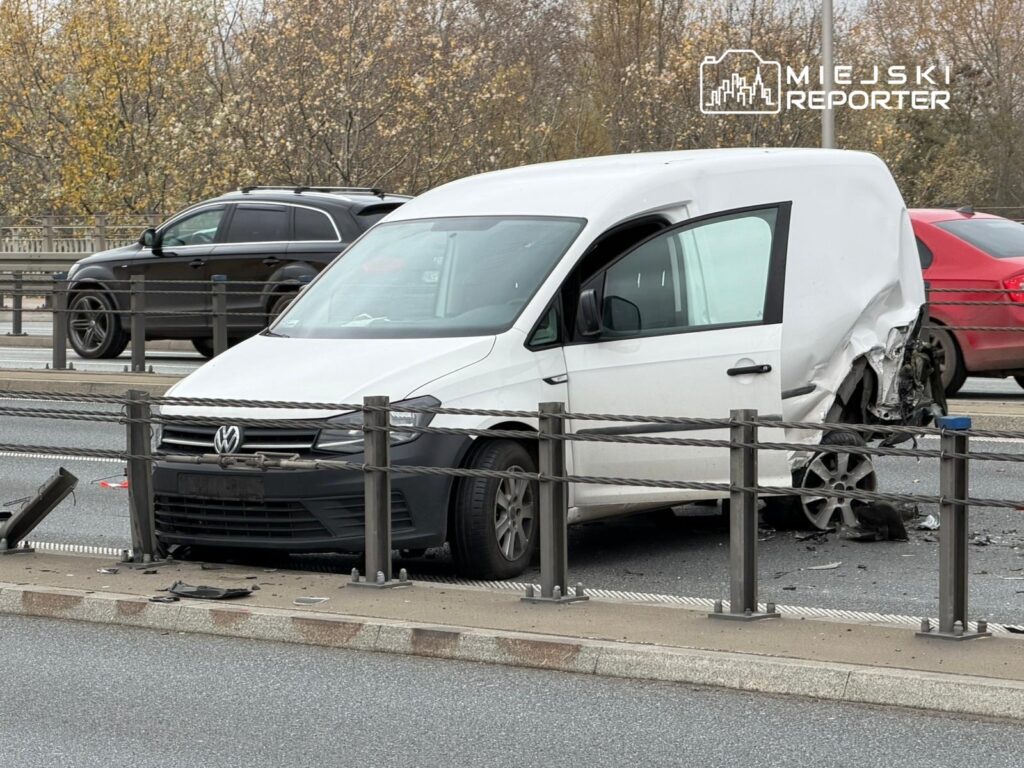 Biały van Volkswagen z uszkodzoną tylną częścią stoi na pasie drogowym, obok metalowego bariery.