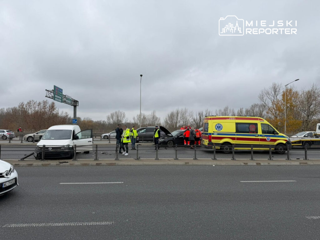Na drodze stoi biały samochód dostawczy z otwartymi drzwiami, a obok niego pracownicy w żółtych kamizelkach zabezpieczają miejsce wypadku. W tle widoczna karetka oraz inne pojazdy.