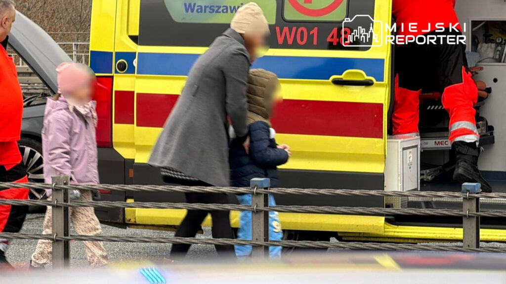 Kobieta w płaszczu trzyma dziecko w kurtce, podchodząc do karetki pogotowia w miejskim otoczeniu.