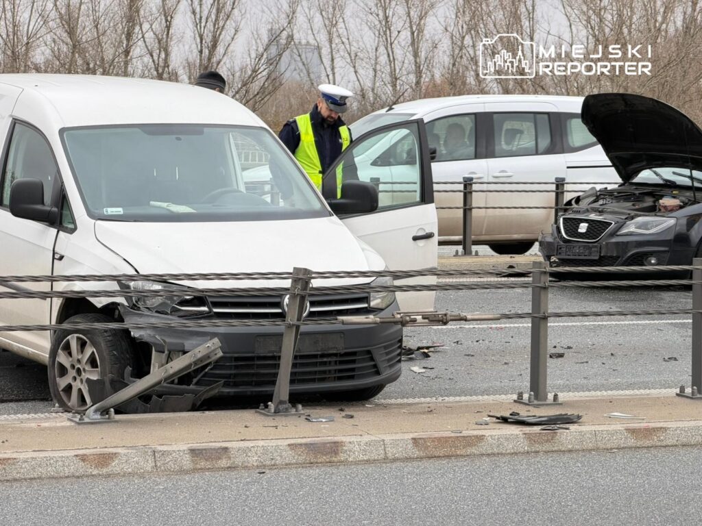 Biały dostawczy samochód z uszkodzoną przednią częścią stoi na drodze, obok policjant w żółtej kamizelce bada sytuację.