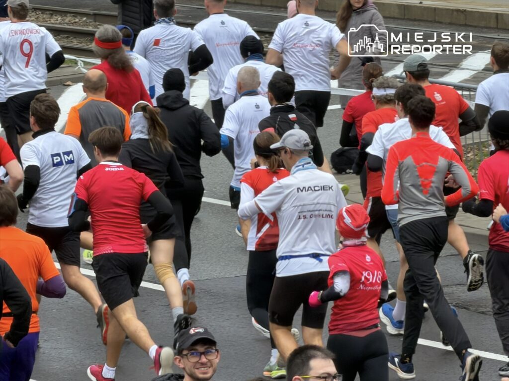 Grupa biegaczy w różnokolorowych koszulkach biegnie ulicą w miejskim otoczeniu podczas wydarzenia sportowego.