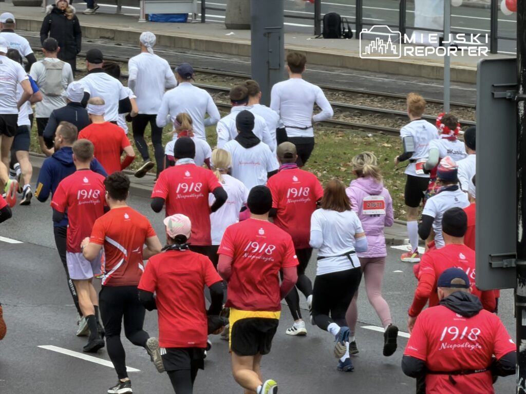 Biegacze w czerwonych koszulkach z napisem "1918 Niepodległa" uczestniczą w biegu ulicami miasta.