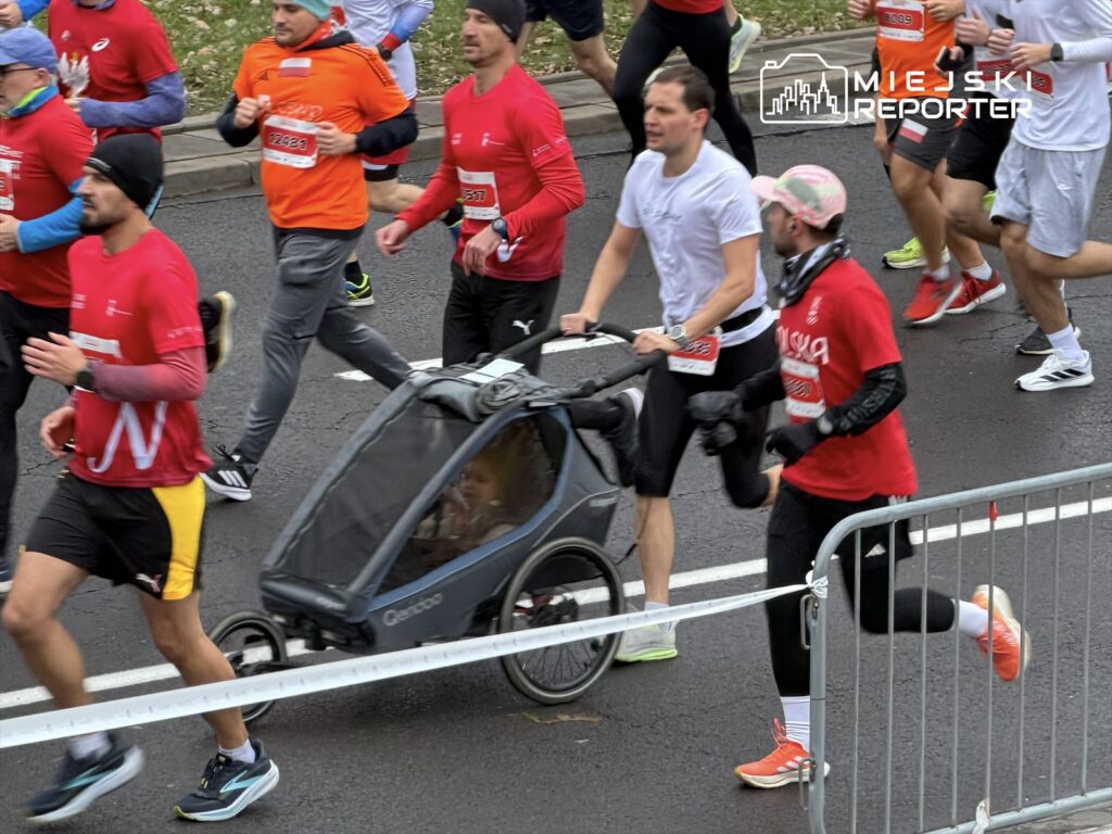 Biegacze w kolorowych strojach uczestniczą w biegu ulicznym, a jeden z nich pcha wózek dziecięcy.