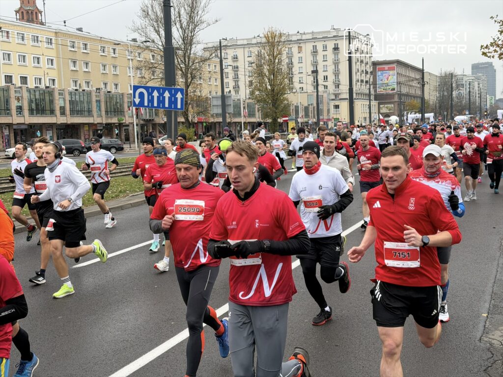 Biegacze w czerwonych koszulkach uczestniczą w wyścigu na ulicach miasta, mijając budynki i znaki drogowe.