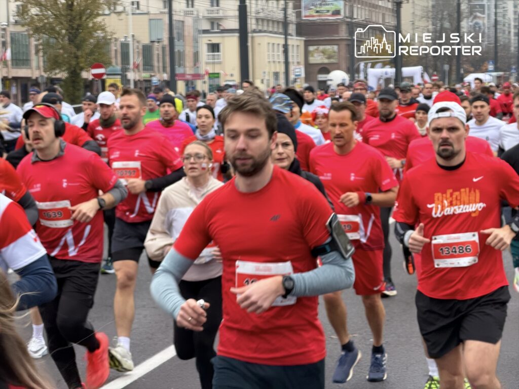 Grupa biegaczy w czerwonych koszulkach uczestniczy w wydarzeniu biegowym na miejskiej ulicy w pochmurny dzień.