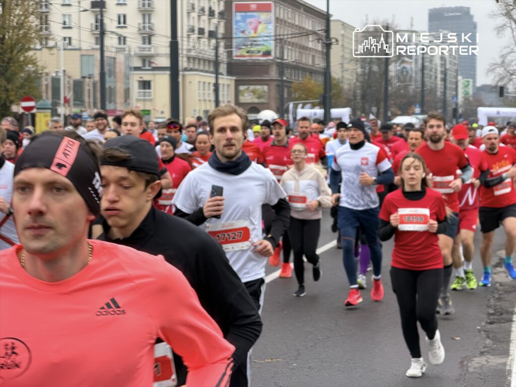 Grupa biegaczy w czerwonych i białych koszulkach uczestniczy w wyścigu ulicznym na zatłoczonej drodze.
