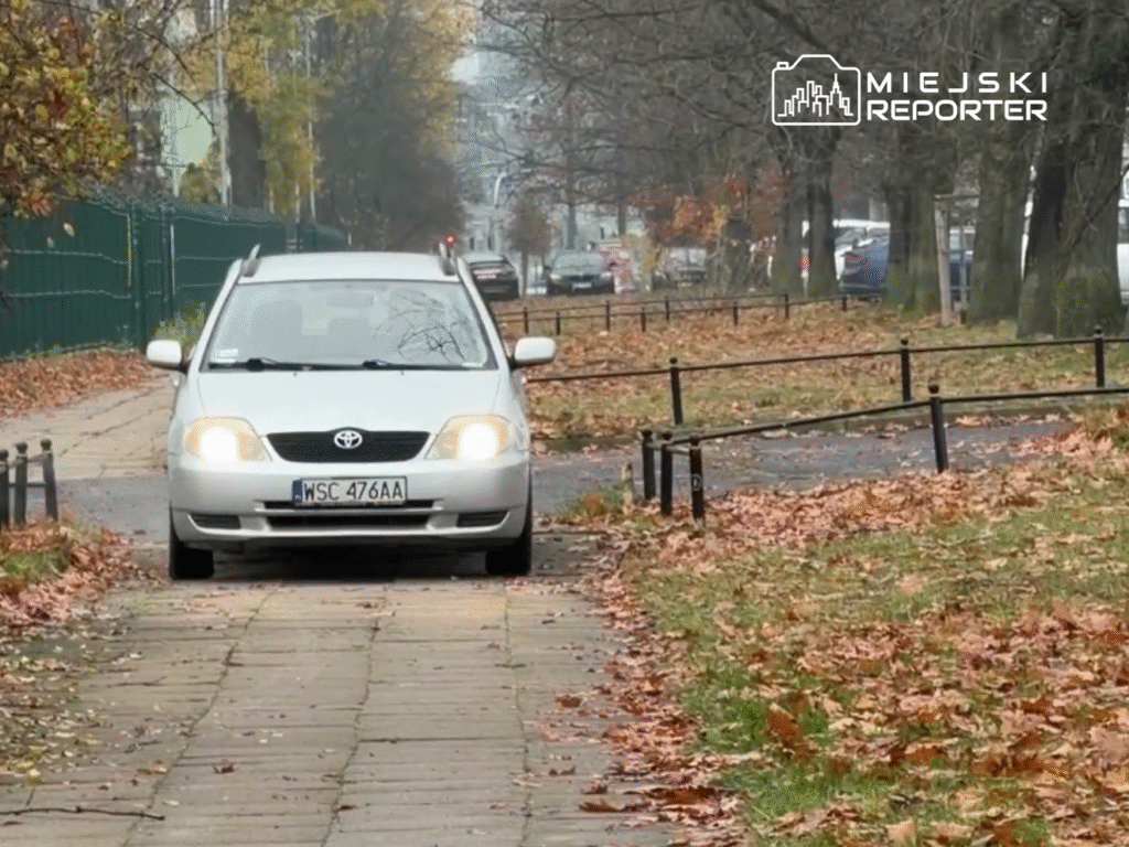 Biały samochód osobowy przejeżdża po chodniku, obok opadłych liści w miejskim otoczeniu.