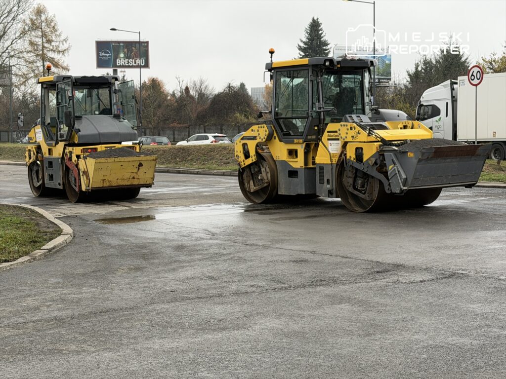 Dwa walce drogowe w żółto-czarnym kolorze stoją na mokrej nawierzchni, w tle widoczne samochody i billboard.