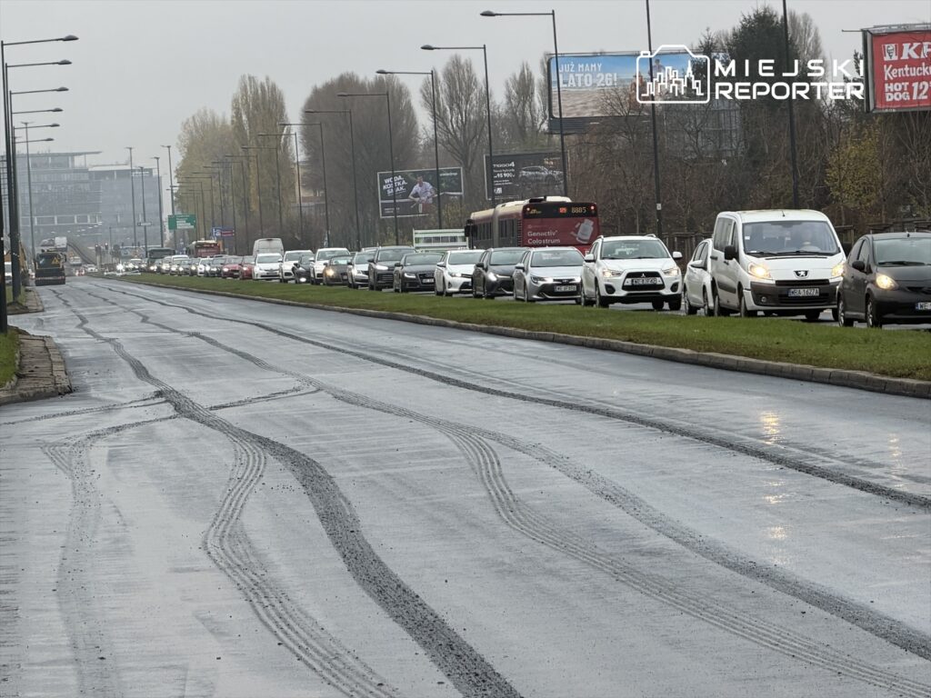 Ruch samochodowy w deszczowy dzień na zatłoczonej ulicy, z widocznymi śladami opon na nawierzchni.