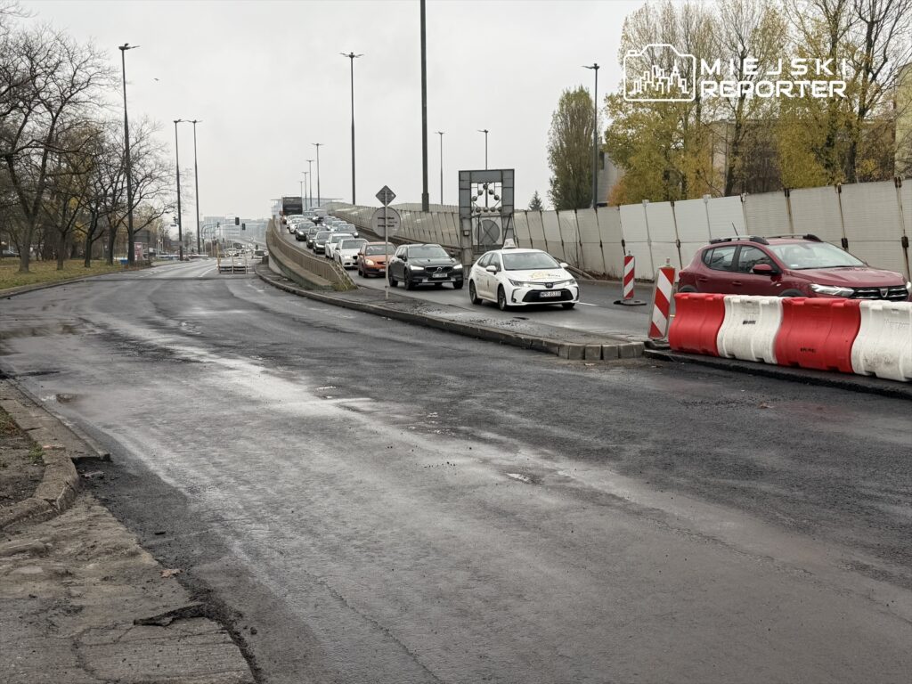 Ruch samochodowy na drodze z wyraźnie widocznymi barierkami drogowymi i oznakowaniem w tle.