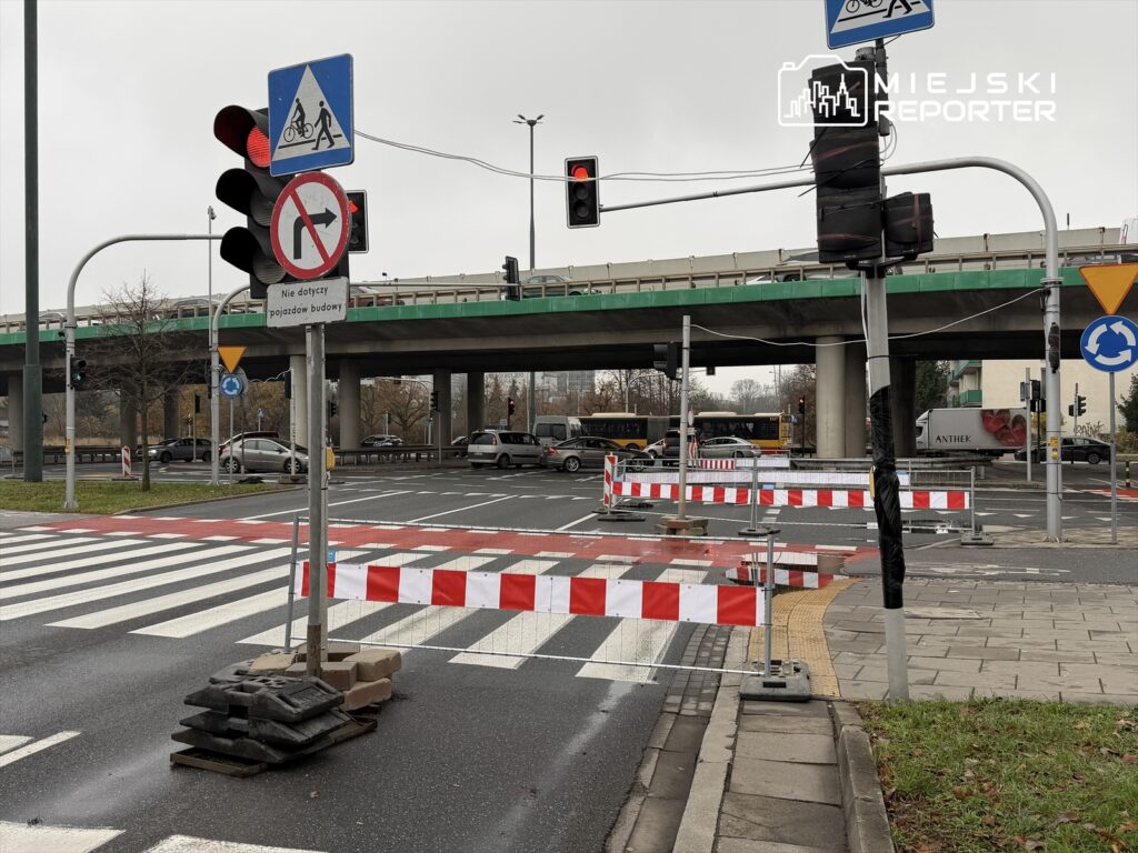 Czerwone światło na sygnalizatorze, oznakowanie drogi, barierki budowlane i znaki zakazu w ruchu w miejskim otoczeniu.