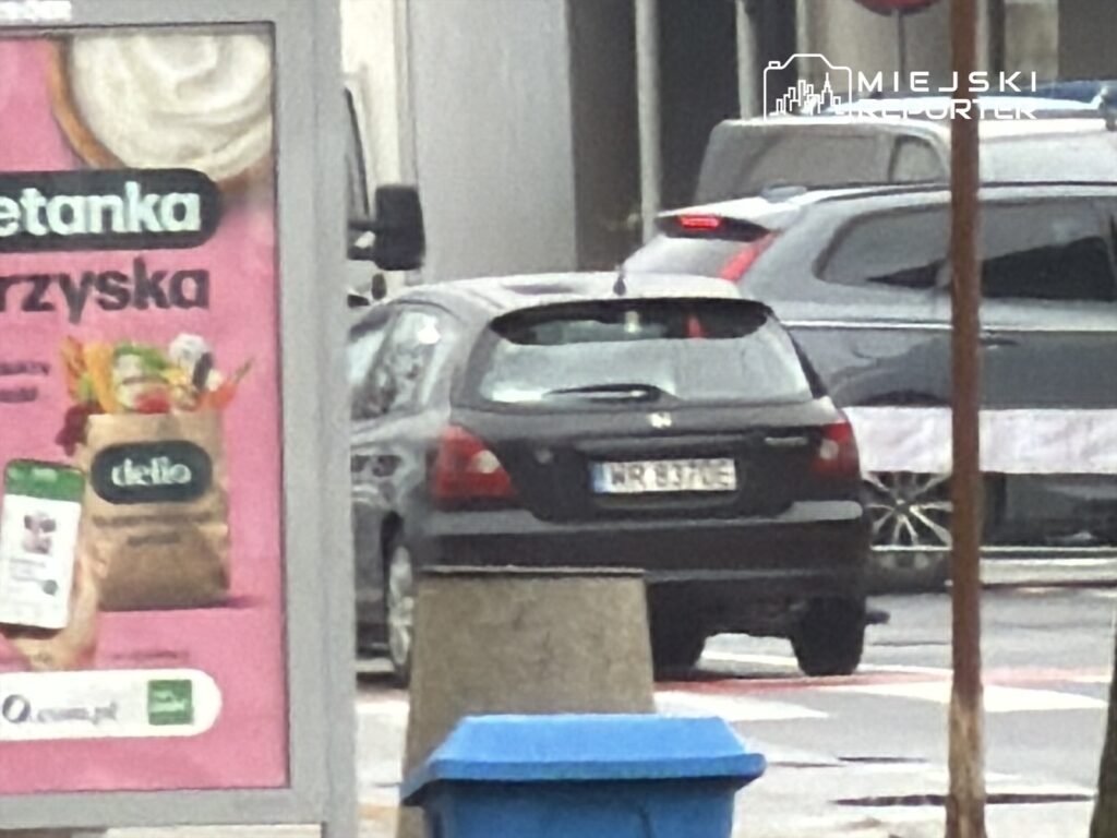 Czarny samochód osobowy zaparkowany obok przystanku, w tle widoczny plakat z ofertą zakupów.