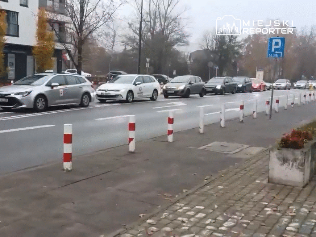 Ruchliwe ulice z samochodami osobowymi i taksówkami, w tle widoczne drzewa i znaki drogowe.
