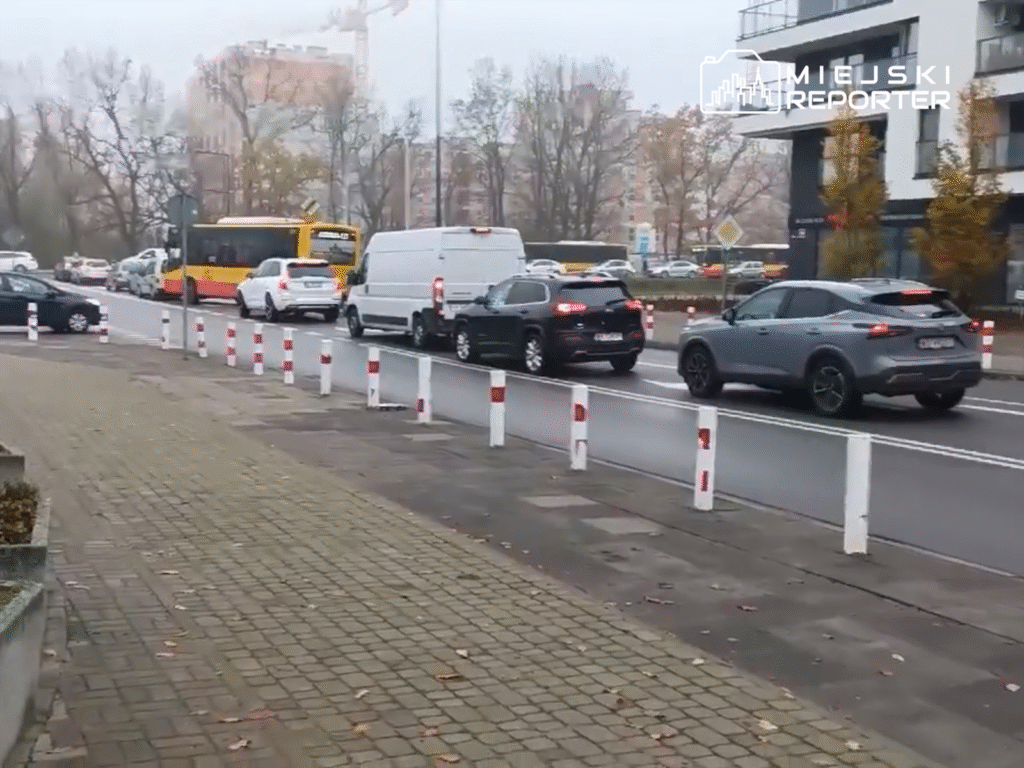 Ruch uliczny z wieloma samochodami i autobusem w tle, w otoczeniu nowoczesnych budynków i drzew.