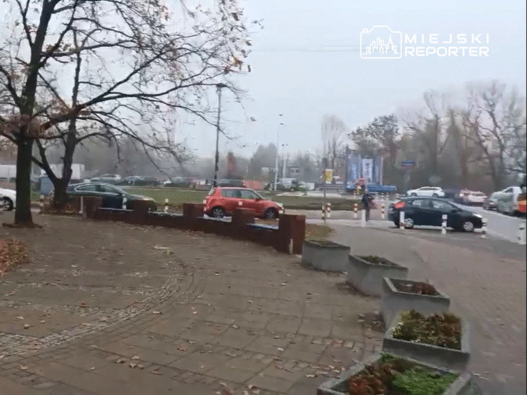 Ruchliwe skrzyżowanie w pochmurny dzień, na pierwszym planie samochody oraz osoby spacerujące po chodniku.