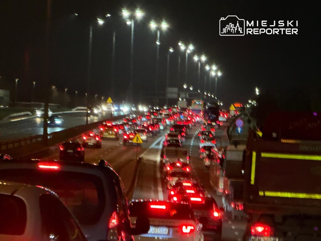 Gęsty ruch samochodowy na autostradzie w nocy, z widocznymi światłami pojazdów i latarniami.