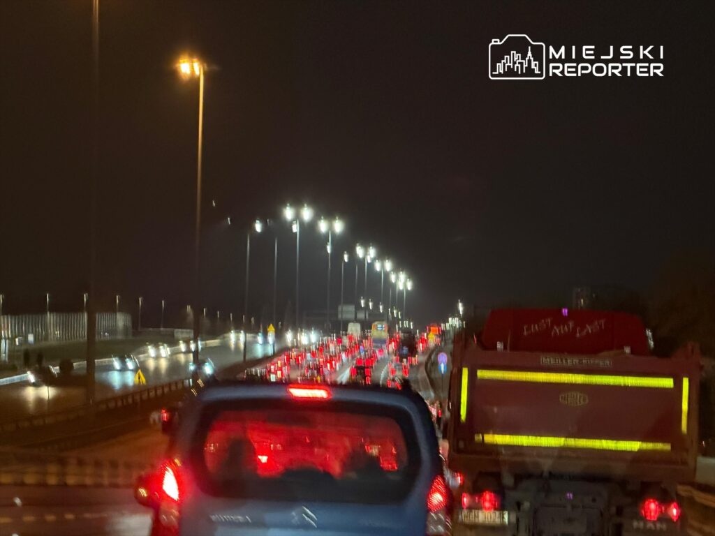 Kolumny samochodów w korku na oświetlonej drodze w nocy, z widocznymi tyłami pojazdów i świateł.