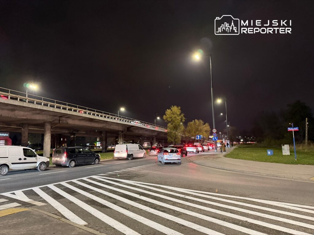 Ruch samochodowy na ulicy z widoczną estakadą, nocą, z latarniami oświetlającymi drogę.
