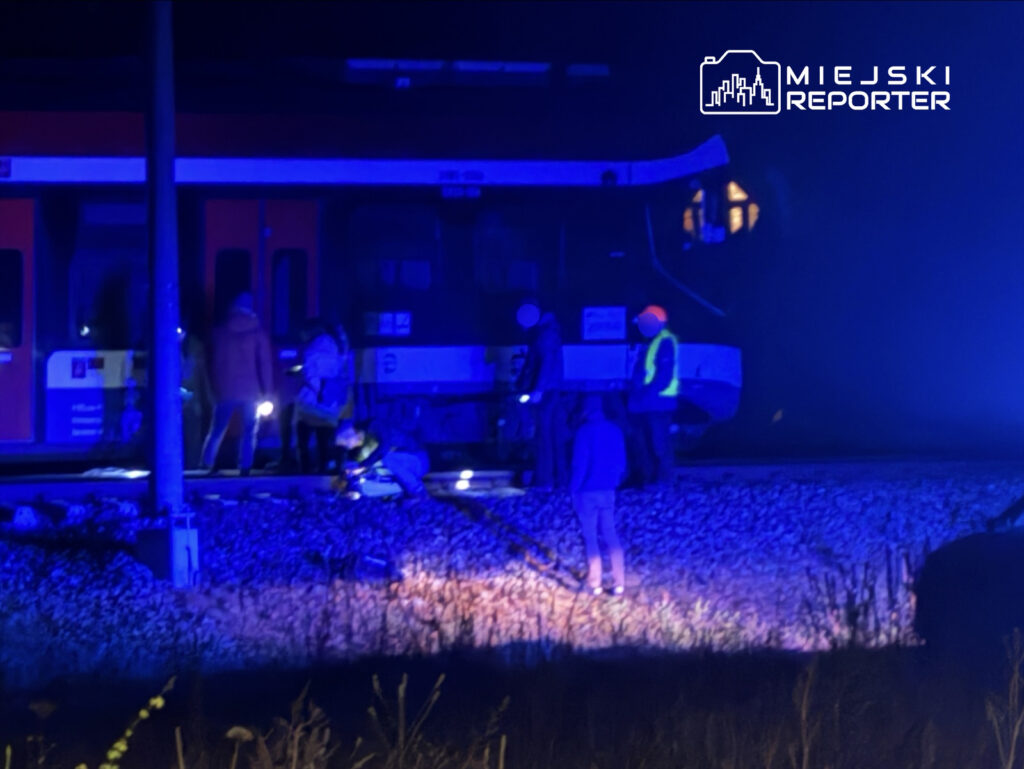 Grupa pracowników w odblaskowych kamizelkach prowadzi prace przy torach kolejowych w nocnym świetle.