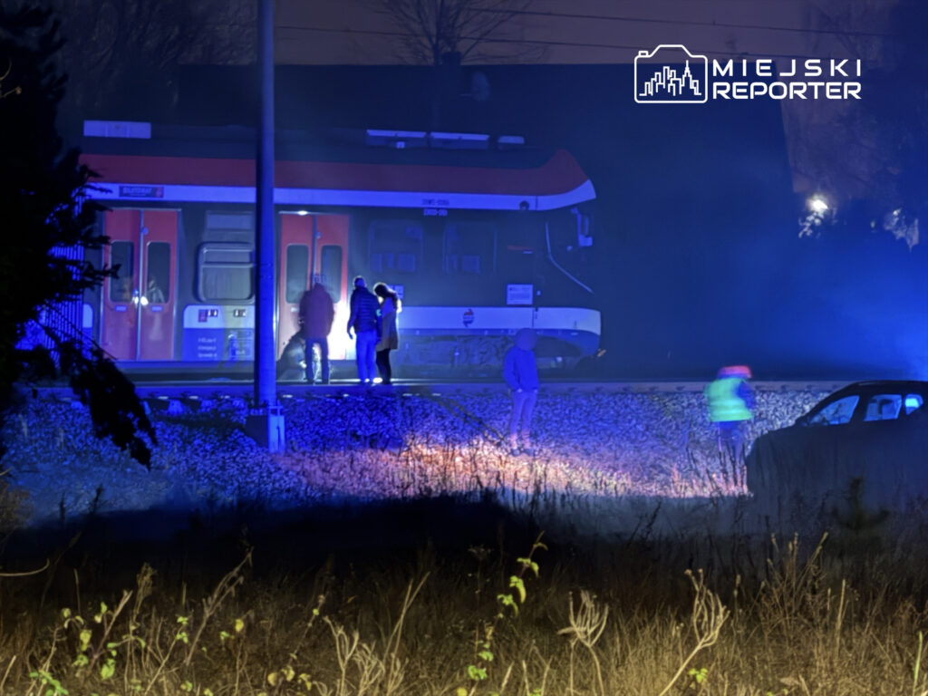 Grupa osób stoi obok torów kolejowych, w pobliżu pociągu, w otoczeniu niebieskiego światła.