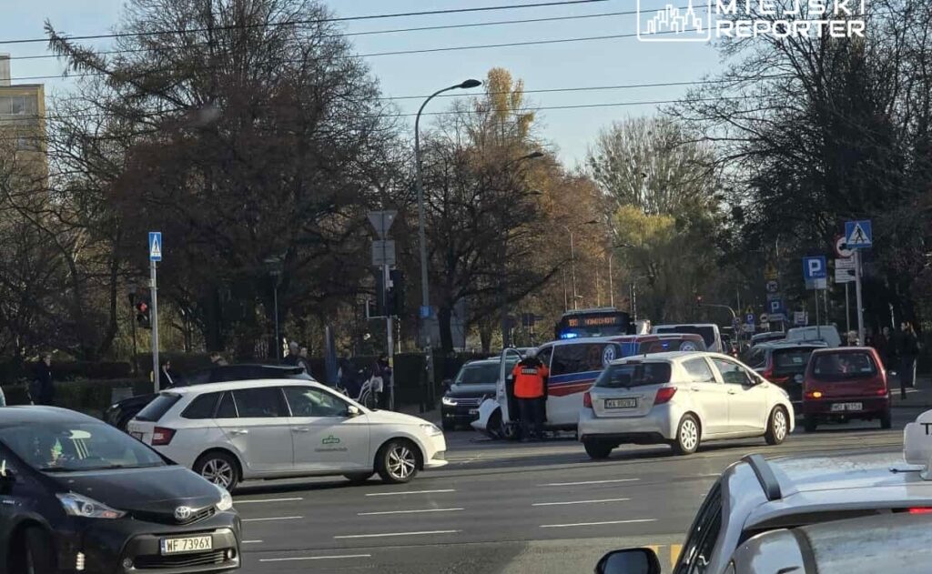 Grupa ludzi stoi obok drogi, podczas gdy karetka i samochody osobowe są zatrzymane w ruchu drogowym.