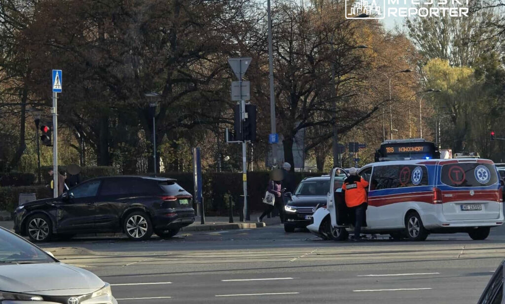 Na skrzyżowaniu stoją dwa uszkodzone samochody, a obok nich karetka pogotowia z personelem medycznym.