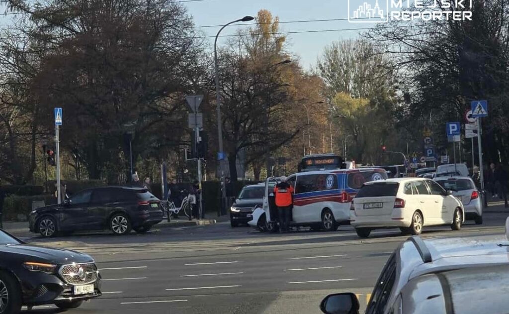 Grupa ludzi obserwująca zdarzenie drogowe, w tle ambulans oraz kilka samochodów na ruchliwej ulicy.